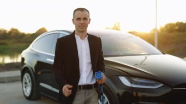 Kafkas işadamının portresi elektrik şarj istasyonunun yanında duruyor kameraya bakıyor ve başparmağını gösteriyor. Erkek elinde şarj olan tip 2 mennekes bir kablo tutuyor. Çevre dostu ulaşım.