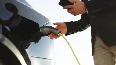 Tanınmayan işadamı elektrik istasyonundan elektrikli arabanın fişini çekiyor. Erkek, akıllı telefon uygulaması kullanarak şarj istasyonunda elektrikli araba yüklüyor. Adam elektrik kablosunu fişten çekiyor..