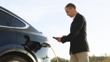 Erkek, güneş ışığında EV aracını şarj ediyor. İşadamı şarj istasyonunda sıfır emisyonlu elektrikli arabanın fişini çekiyor. Adam akıllı telefondaki uygulamayı kullanarak şarj kablosunu elektrikli araca bağladı.