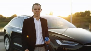 Kendine güvenen genç profesyonel iş adamı lider kazanan çalışan kameraya bakıyor. Dışarıdaki elektrikli arabanın yanındaki şarj istasyonunda el işaretini gösteriyor..