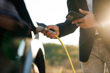 Erkek, güneş ışığında EV aracını şarj ediyor. İşadamı şarj istasyonunda sıfır emisyonlu elektrikli arabanın fişini çekiyor. Adam akıllı telefondaki uygulamayı kullanarak şarj kablosunu elektrikli araca bağladı.