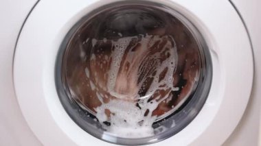 Washing clothing in domestic washing machine in home. Close-up video of spinning drum washing machine. White washing machine washes dirty clothes.