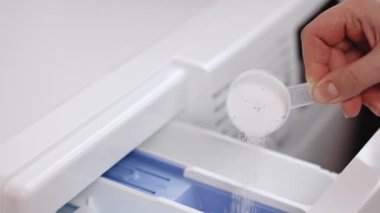 Woman carefully poured laundry detergent into compartment of washing machine in bathroom. With white measuring spoon person fills washing machine with powder.