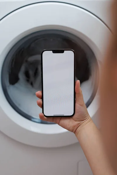 Woman holds a mobile phone with a white screen in her hands. Girl use mobile phone control washer at home. Washing machine, smart home. Smartphone device. Smart washing machine concept.