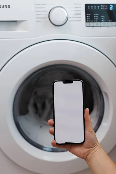 Woman using digital smart phone on laundry during waiting smart washing machine working, self-service laundry facility, setting application for washing on smartphone white screen.