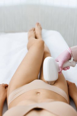 Modern salonda güzellik lazeri cilt bakımı sırasında bayan müşterinin bikini bölgesinde lazer tüy temizleme aparatı kullanan eldivenli anonim güzellik uzmanı. Güzellik kliniğinde lazer epilasyonu.