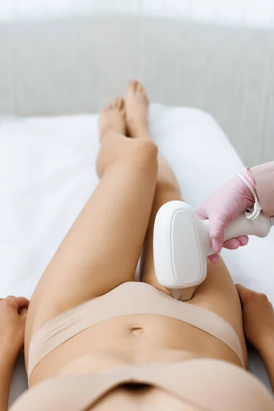 Anonymous beautician in gloves using laser hair removal apparatus on bikini zone of female client during beauty laser skin care procedure on stomach in modern salon. Laser epilation at beauty clinic.
