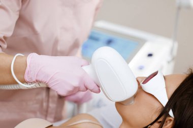 Lazer bıyık aldırma. Yüz kılları alınıyor. Gözlüklü kadın, lazer tüy çıkarma prosedürü uyguluyor. Tüy dökümü için bir cihaz. Cilt bakımı, kozmetik prosedürleri..