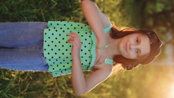 ハッピーかわいい少女の垂直ビデオは 屋外のカメラで笑顔で腕を横切りました 愛らしいプリスクールの幼稚園の子供の肖像画 日没でぼやけた背景の上に公園に立っている笑顔 — ストック動画