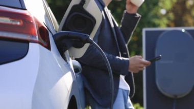 Şarj istasyonundan elektrikli bir araca şarj oluyor. Elektrikli bir arabanın yanında duran tanınmayan adam şarj oluyor ve akıllı telefondan zaman ayarları yapıyor..