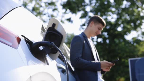 充電している電気自動車の近くに立っている豪華な慎重なビジネスマンは スマートフォンで時間を調整します 充電ステーションから電気自動車を充電するプラグ — ストック動画