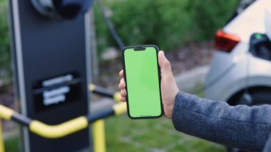 EV vagonunu şarj edebilmek için şarj istasyonuna cep telefonuyla bağlanmak. Arka plandaki elektrikli araç şarja takılıyor. Cep telefonundaki krom anahtarı taklit et.