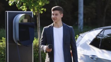 Kendine güvenen genç profesyonel iş adamı lider kazanan çalışan kameraya bakıyor. Dışarıdaki elektrikli arabanın yanındaki şarj istasyonunda el işaretini gösteriyor..
