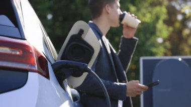 Elektrikli arabanın yanında duran beyaz adam şarj oluyor ve akıllı telefondan zaman ayarları yapıyor. Bir elektrikli arabayı şarj istasyonundan şarj etmek.