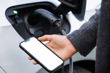 Elektrikli araç şarj istasyonunda akıllı telefon tutan adam, yakın plan. Mobil uygulamalı elektrikli araba şarj istasyonu.