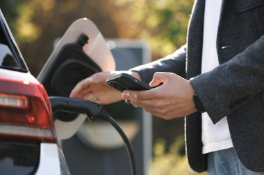 Businessman charging electric car at outdoor charging station Unrecognizable man unplugging electric car from charging station. Male unplugging in power cord to electric car using app on smartphone.