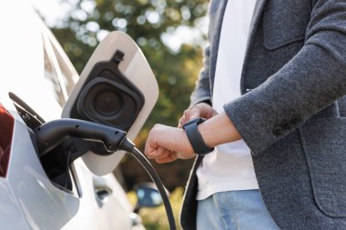 Male hand unplugs power connector into EV car and charges batteries, uses smart watch to disconnect charging. Man disconnects charging cable to to electric vehicle using smartwatch.