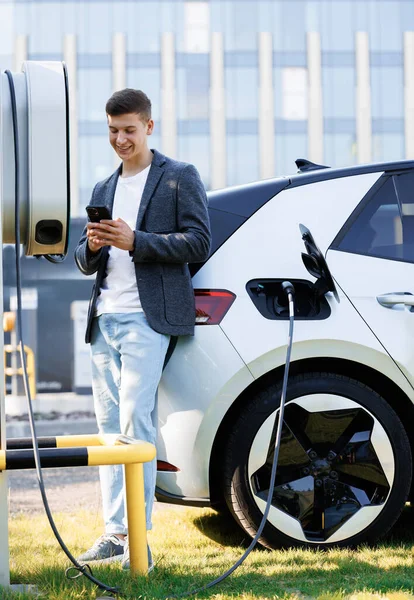 Genç Adam Şarj Istasyonunda Elektrikli Arabanın Yanında Akıllı Telefon Kullanıyor — Stok fotoğraf