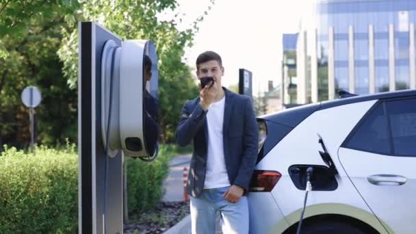 商人使用智能手机语音信息和等待电源连接到电动汽车充电的汽车 从充电站给电动车充电 — 图库视频影像