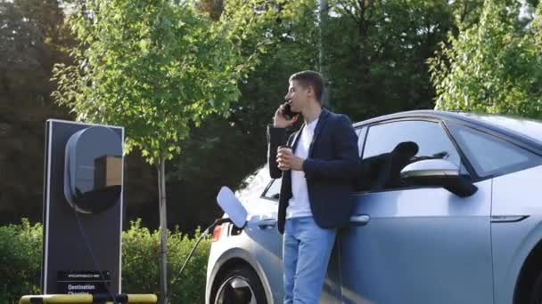 ウクライナのリヴィウス 2023年8月19日 電気自動車の充電バッテリー中にモバイル会話を持つハンサムなビジネスマン フォルクスワーゲンId3電気自動車が充電ステーションに差し込まれ充電 — ストック動画