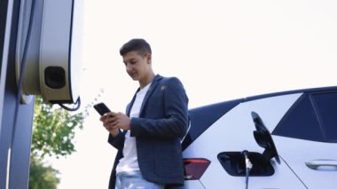 Beyaz adam akıllı telefon kullanıyor ve elektrikli araçlarla bağlantı kuruyor. Arabadaki aküyü şarj etmek için. Bir elektrikli arabayı şarj istasyonundan şarj etmek.