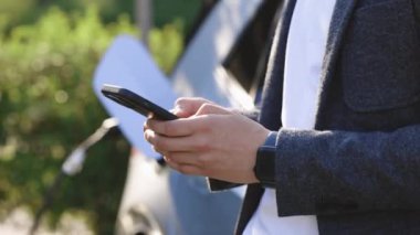 Elektrikli araç şarj istasyonunda akıllı telefon tutan adam, yakın plan. Elektrikli araba şarj oluyor, iş adamı arkada duruyor ve bekliyor..
