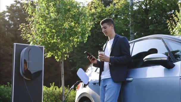 Καυκάσιος Άντρας Στέκεται Κοντά Ένα Ηλεκτρικό Αυτοκίνητο Που Φορτίζει Και — Αρχείο Βίντεο