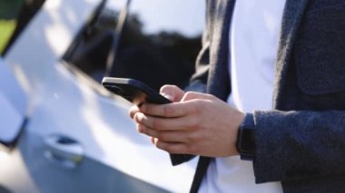 Genç adam şarj istasyonunda elektrikli arabanın yanında akıllı telefon kullanıyor. Elektrikli araç şarj istasyonunda akıllı telefon tutan adam. İş adamı arka planda duruyor ve bekliyor.