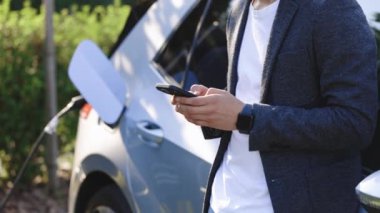 Akıllı telefon kullanan işadamı ve beklemede olan güç kaynağı arabadaki bataryayı şarj etmek için elektrikli araçlara bağlanıyor. Bir elektrikli arabayı şarj istasyonundan şarj etmek.