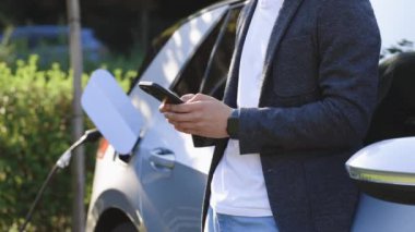 Şarj istasyonundan bir elektrikli arabayı şarj etmek. Beyaz adam akıllı telefon kullanıyor ve güç kaynağı bekliyor. Arabanın aküsünü şarj etmek için elektrikli araçlara bağlanıyor..