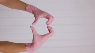 Pembe lateks eldivenli birinin dikey videosu kalp hareketlerini beyaz arka planda, yakın planda gösteriyor. Kalp şeklinde eldiven takan eller tıbbi ekibe sevgiyi ya da desteği simgeler..