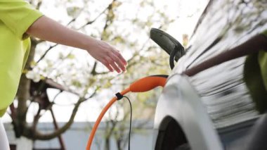 Kadın elleri elektrikli otomobilinin fişini çekiyor ve evinin önündeki kapağı kapatıyor. Beyaz bir elektrikli arabanın elektrik kablosunun fişini çeken kadın.