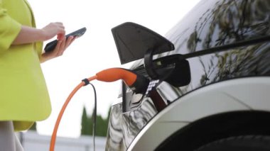 Elektrikli araba kablosunun fişini çeken kadın. Kız cep telefonu akıllı telefonunda uygulamayla şarj olmayı kesti. Çevre dostu nakliye aracı. Kadın eli elektrik kablosunu koparıp elektrikli arabayı şarj eder..