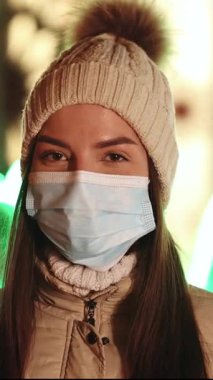 Süslü Xmas şehrinde duran, beyaz, tıbbi maskeli güzel bir kadının dikey video portresi. Kalabalık covid-19 'u hasta etti. Salgın koronavirüs. Salgın grip korona virüsü