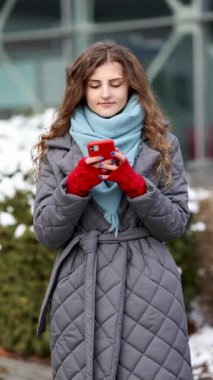 Elinde telefon tutan, kışın giydiği kıvırcık kadının dikey video portresi güzel mobil haber teklifini kutluyor. Heyecanlı kadın kazanan evet diye bağırıyor. Cep telefonuna bakarak mutlu bir başarı elde ediyor..
