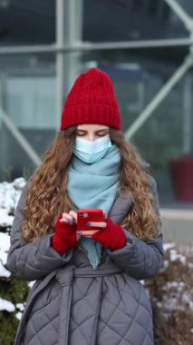 Neşeli beyaz kadın portresinin dikey videosu. Maskeli güzel, kıvırcık kadın sokakta dikilirken akıllı telefondan bakıyor. Neşeli genç bir kadın cep telefonuyla mesaj atıyor. Dikey Ekran Yönelimi 4k