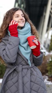 Şehir caddesinde yürürken cep telefonuyla konuşan kıvırcık kızın dikey videosu. Kıvırcık saçlı kadın akıllı telefonunu kullanıyor ve elinde kahve ile kırmızı kağıt bardak tutuyor. Yürüyen ve konuşan kadın