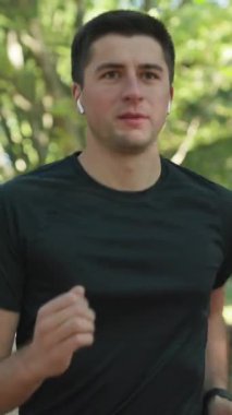 Bluetooth kulaklık ve akıllı saat uygulaması kullanarak sağlıklı ve aktif yaşam tarzının tadını çıkaran bir adamın şehir parkında koştuğunu gösteren dikey video. Erkek koşucu yaz güneşinde kardiyo egzersizi yapıyor.