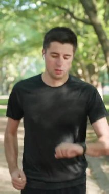 Sağlıklı ve aktif yaşam tarzının dikey videosu. Uzaklık uygulaması. Parkta boş bir yolda koşan sporcu dışarıda eğitim görürken kablosuz kulaklık kullanarak internet üzerinden akıllı saat uyarısını kontrol ediyor..