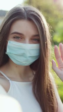 Maskeli genç kadın vlogger kameraya bakarak el sallarken çekilmiş dikey bir video. Çevrimiçi olarak arkadaşlarıyla iletişim kuran kız video görüşmesi yapıyor. Konuşan yaşam tarzı vlog iş görüşmesi.
