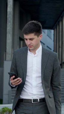 Takım Elbiseli İşadamı 'nın dikey Format Videosu Ofis Yakınındaki Bir Sokakta Akıllı Telefon Kullanıyor. Ofisteki diğer insanlar yanımızdan geçiyor. Gülümsüyor ve başarılı görünüyor. Aygıtında Web 'i tarıyor