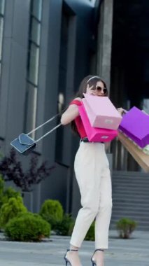 Kırmızı ve bej pantolonlu güzel bir kadının dikey videosu renkli alışveriş torbalarıyla bir binanın önünde duruyor, gülümsüyor ve başka tarafa bakıyor. Tam boy vücut boyutu görünümü.