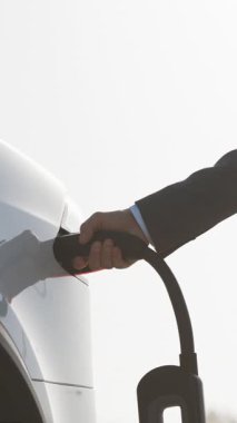 Elektrikli arabanın fişini çeken iş adamının dikey videosu. Lüks beyaz elektrikli araba dolu. Elektrik güç dolumu yarışması ve kablo bağlantısı koptu. Ekoloji alternatif enerjisini gelecek sürdürülebilir olarak kaydet.