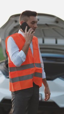 Adamın telefonda konuştuğu ve arabanın motoruna baktığı dikey video. Genç adam akıllı telefon kullanıyor, bozuk arabanın yanında duruyor kaputu açıyor, araba yardım servisini arıyor, tamirata yardım ediyor.