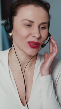 Telefon merkezi çalışanının dikey format videosu. Kulaklıkla arayan müşteriyle konuşmaktan memnun. Mutlu çağrı merkezi ajanı, parlak bir ofiste kulaklıklarıyla konuşuyor..