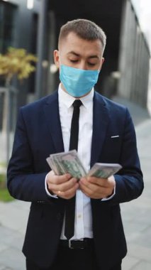 Dolarları elinde tutan ve hesaplayan yakışıklı bir adamın dikey format videosu. Koruyucu maskeli neşeli, zengin, yakışıklı iş adamı iş merkezinin arka planında para sayıyor..