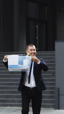 Mutlu iş adamının iş merkezinden ayrılıp dans ettiği kutularla sokakta yürüdüğü dikey video. Ofis çalışanı işi bırakıp kutlama yapıyor. Neşeli adamı kovdum. Harika bir gün, iyi bir ruh hali..
