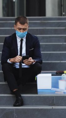 Dışarıdaki merdivenlerde kişisel eşyalarıyla oturan tıbbi maskeli kovulmuş bir adamın dikey videosu. Yoksul erkek iş yerini kaybetti ve akıllı telefondan iş arıyor. Pandemik ekonomik kriz.