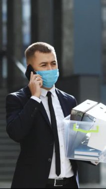 Kovulmuş bir iş adamının modern ofis binasının yakınındaki kutudaki eşyalarla telefonda konuştuğu dikey video. Koruyucu yüz maskesi takan iş adamı şehir merkezindeki sesli mesajları göndermek için akıllı telefon kullanıyor..