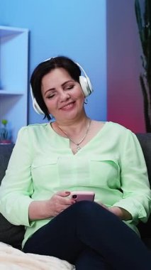 Teknolojinin Dikey Videosu, Yaşlılar. Evde Müzik Dinleyen Kulaklıklı Mutlu Son Sınıf Kadınları. Kablosuz kulaklık takan yaşlı bir kadın favori bir şarkıyı dinliyor ve gülümsüyor..